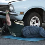 A mechanic working under a classic car on the pavement during the day — SSK Mechanics guide to Peugeot 208 Brake Pads, Discs and Calipers Guide