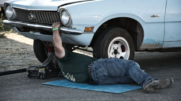 A mechanic working under a classic car on the pavement during the day — SSK Mechanics guide to Peugeot 208 Brake Pads, Discs and Calipers Guide
