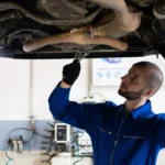 A mechanic in blue overalls inspects the exhaust system of a car in a workshop — SSK Mechanics guide to What Does White Smoke From the Exhaust Mean