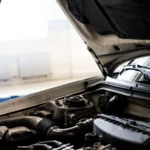 Detailed image of a car engine underneath the open hood in a well-lit indoor garage — SSK Mechanics guide to What Does Blue Smoke From the Exhaust Mean
