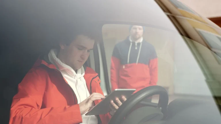 A man in a red jacket uses a tablet inside a car as seen through the windshield — SSK Mechanics guide to What Does It Really Cost to Own a Car? The Main Factors Explained