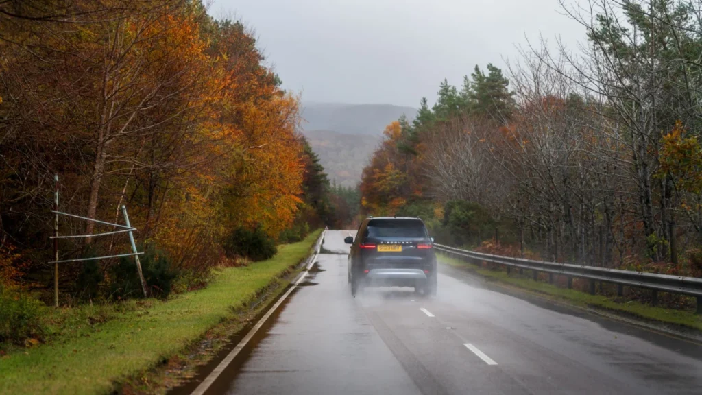 Car driving down a wet road surrounded by colorful autumn trees under a cloudy sky — SSK Mechanics guide to Driving in Bradford & West Yorkshire: What Local Roads Do to…