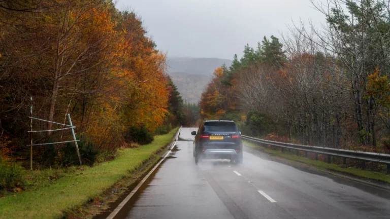 Car driving down a wet road surrounded by colorful autumn trees under a cloudy sky — SSK Mechanics guide to Driving in Bradford & West Yorkshire: What Local Roads Do to…