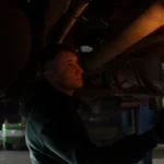 Mechanic working in dimly lit garage, inspecting car's undercarriage with a portable light source — SSK Mechanics guide to Spen Valley Roads: Pothole Damage Around Cleckheaton, Low…