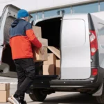 Man in colorful jacket loading cardboard boxes into a van outside an office building — SSK Mechanics guide to Why Sole Traders Are Switching to On-Site Van Servicing