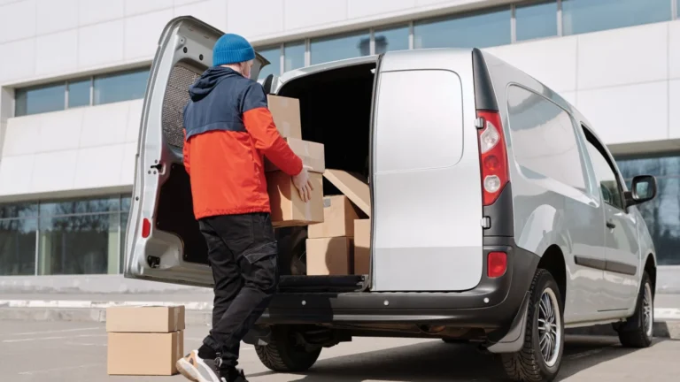 Man in colorful jacket loading cardboard boxes into a van outside an office building — SSK Mechanics guide to Why Sole Traders Are Switching to On-Site Van Servicing