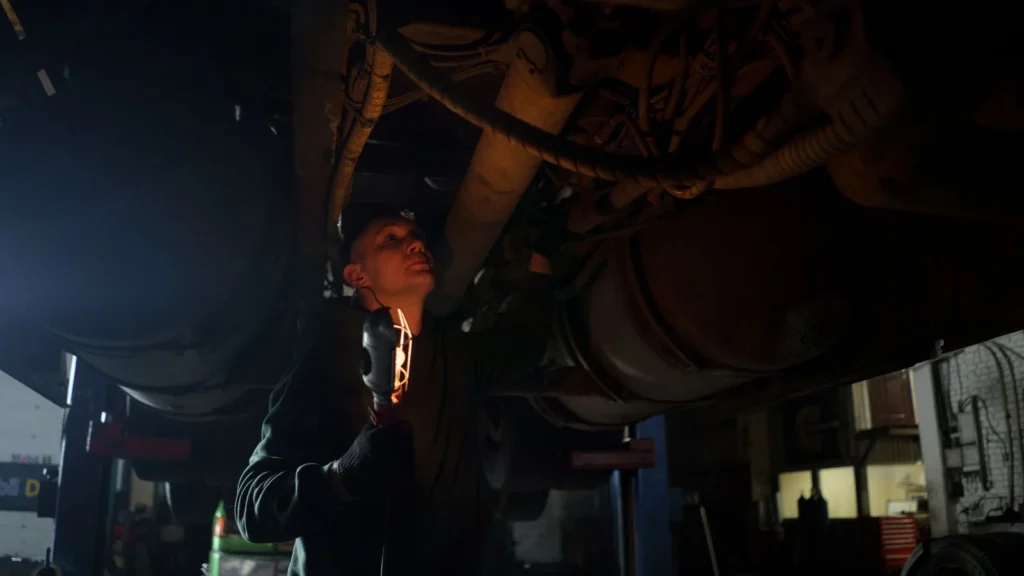 Mechanic examines vehicle underbody in dimly lit workshop, holding inspection light — SSK Mechanics guide to Why You Should Never Jack a Car Up Without Axle Stands