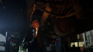 Mechanic examines vehicle underbody in dimly lit workshop, holding inspection light — SSK Mechanics guide to Why You Should Never Jack a Car Up Without Axle Stands
