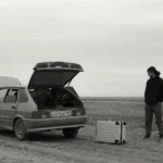 A man stands by a broken down car on a dirt road with luggage in a desolate, rural setting — SSK Mechanics guide to Essential Tools Every UK Driver Should Keep in Their Car