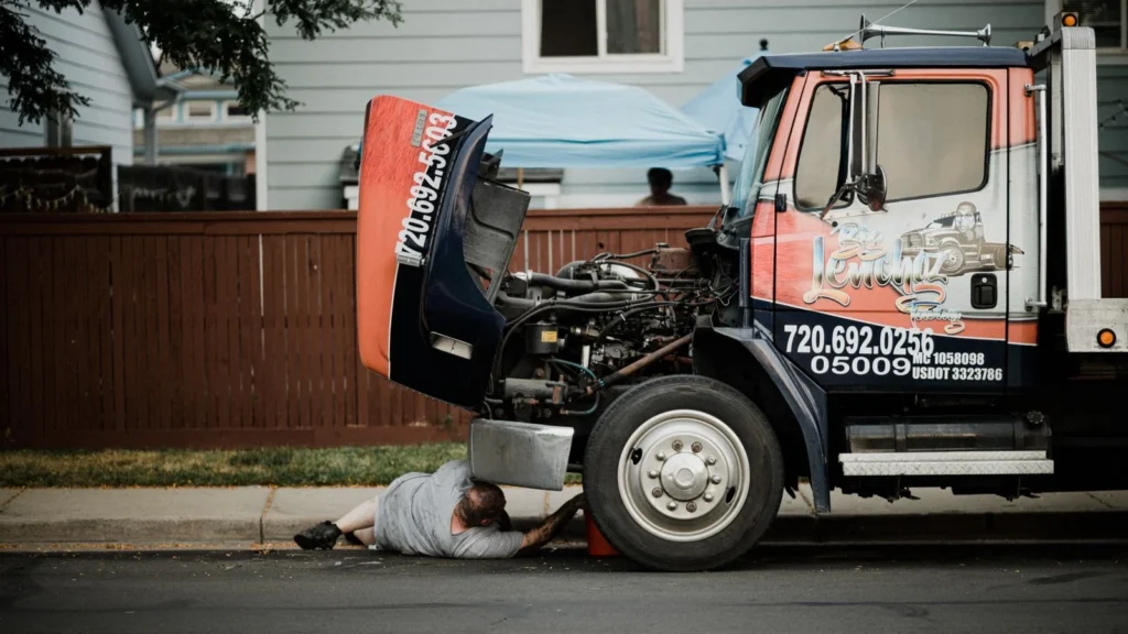 A mechanic works under a truck on a neighborhood street, highlighting vehicle maintenance — SSK Mechanics guide to Kwik Fit Bradford vs a Local Mobile Mechanic: Which Should…