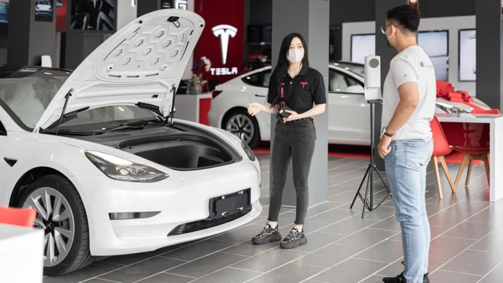 A customer talks with a sales representative about a Tesla Model 3 in a car dealership,… — SSK Mechanics guide to Main Dealer EV Servicing vs Independent Mobile EV…