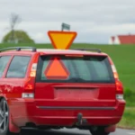 A red car drives on a countryside road in Jönköping, Sweden with a rural landscape — SSK Mechanics guide to Halfords Summer Car Check vs SSK Mechanics: What Each…