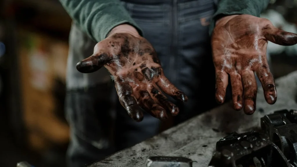Close-up of a mechanic's dirty hands in a garage workspace with grease and spare parts — SSK Mechanics guide to AA/RAC Seasonal Vehicle Checks vs a Local Mobile Mechanic's…